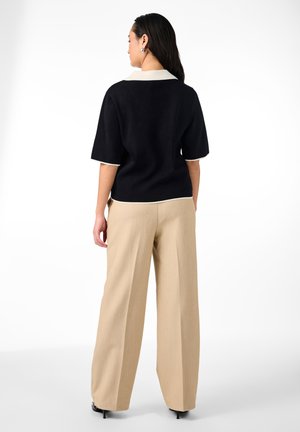 Femme debout de dos portant un haut noir à manches courtes avec un col blanc et un pantalon ample beige avec des talons noirs.
