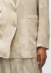 Blazer beige à motif floral avec boutons assortis et grande poche avant, porté avec une jupe assortie et une main reposant à côté.