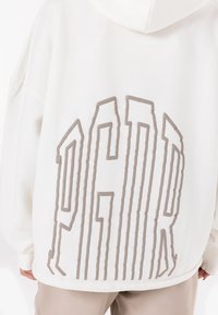 Veľká biela mikina s kapucňou vyrobená z mäkkej textílie, na chrbte s veľkým vyvýšeným béžovým logom "PGHR". S kapucňou a s rebrovanými manžetami.