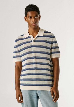 Young man wearing a beige and blue striped short-sleeve knit polo shirt and light blue jeans, standing against a plain background.