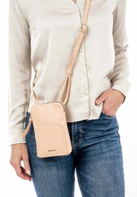 Pfirsichfarbene Leder umhänge Tasche mit glatter Textur, Reißverschluss, Fronttasche und verstellbarem Tragegurt, getragen zu einem hellen Shirt und Denim.