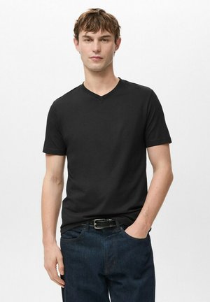 Junger Mann steht mit Hand in der Tasche, trägt ein schwarzes V-Ausschnitt-T-Shirt und dunkle blaue Jeans vor weißem Hintergrund.