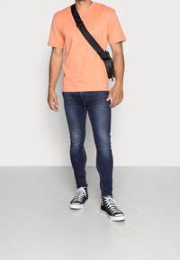 Hombre con camiseta melocotón, jeans ajustados oscuros, zapatillas altas negras y bolso bandolera con correa negra sobre un fondo liso.