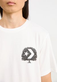 Camiseta de algodón blanca con un estampado gráfico negro de un árbol con una estrella y una flecha dentro del tronco, cuello redondo y mangas cortas.