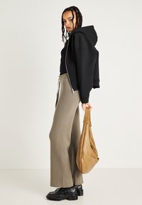 Crna zip-up hoodie, rebrasti maslinasto zeleni široki hlače, crne robustne cipele i smeđa kožna torba preko ramena. Pleteni frizura upotpunjuje izgled.