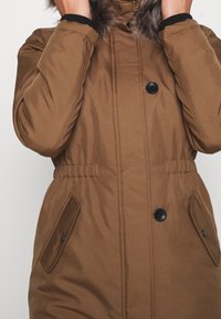 Brown insulated jacket with a fur-lined hood, featuring a buttoned front, elasticized waist, and side pockets. Smooth texture, warm design.