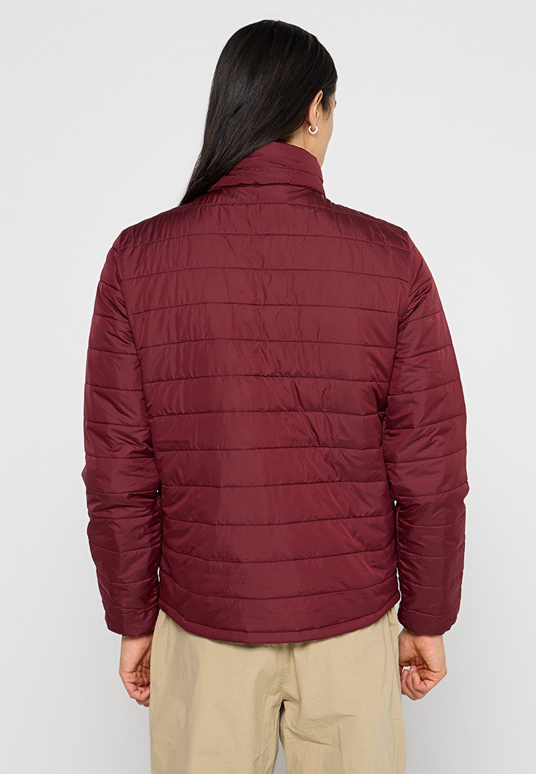 Persona con lunghi capelli scuri che indossa una giacca trapuntata bordeaux e pantaloni beige, vista da dietro su uno sfondo semplice.