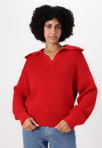 Rød strikket genser med vid krage og løse ermer, med ribbestrukturen og en liten logo på brystet. Kombinert med blå jeans.