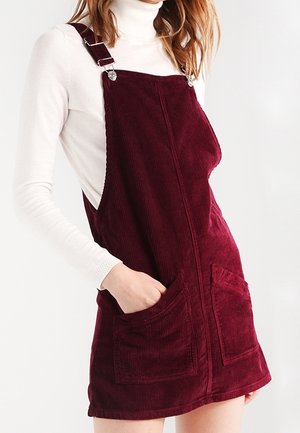 Kvinde iført en hvid rullekrave og en bordeaux kordfløjls-foret kjole med lommer foran, hånden hvilende i en af lommerne.