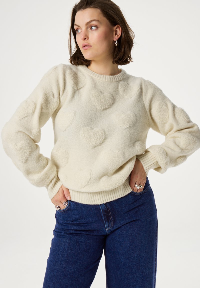 Romantische gebreide crème trui met verhoogde hartpatronen, ronde halslijn en ribgebreide manchetten. Gecombineerd met donkerblauwe jeans.