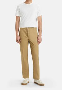 Chinos en coton beige, coupe droite avec une poche avant, associés à un t-shirt blanc uni et des baskets blanches avec des semelles marron clair.