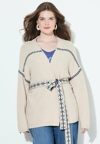 Beige stickad kardigan med långa ärmar, med mörkblå detaljer och ett mönstrat bälte. Bärs över en blå topp, kombinerad med blå denim.
