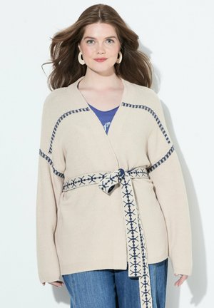 Beige neulecardigan, jossa on pitkät hihat, ja johon on lisätty laivastonsinisiä yksityiskohtia sekä kuvioitu vyö. Päälläsininen toppi, parina siniset farkut.