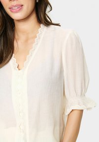 Leichtes, cremefarbenes Blusenshirt mit einem gewellten Ausschnitt, dekorativer Knopfleiste und Rüschenärmeln mit Spitzenakzenten.
