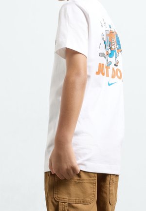 Kind draagt een wit T-shirt met cartoonafbeelding en tekst, en bruine cargoshorts, zijaanzicht tegen een effen achtergrond.