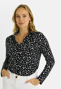 Camisa negra de manga larga con patrón abstracto de puntos en blanco y dorado, con botones en la parte delantera y un cuello, combinada con pantalones blancos.