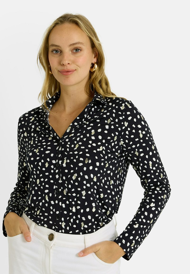 Camisa negra de manga larga con patrón abstracto de puntos en blanco y dorado, con botones en la parte delantera y un cuello, combinada con pantalones blancos.