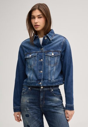 Jonge vrouw die een blauwe spijkerjasje met knoopsluitingen en versleten blauwe jeans draagt tegen een effen achtergrond.