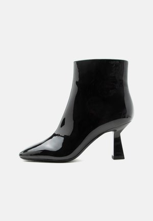 Zwarte glanzende lakleren enkellaars met spitse neus en uitlopende middelhoge hak op witte achtergrond.