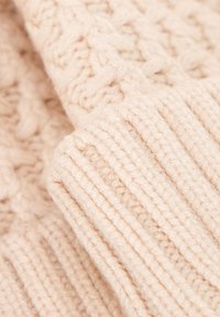 Dettaglio di maglione lavorato a maglia color beige chiaro con motivi strutturati e bordi a coste, realizzato con filato morbido, caratterizzato da un design accogliente e spesso.