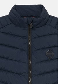 Veste matelassée bleu marine avec col haut, zip central noir et patch du logo Hackett sur la poitrine.