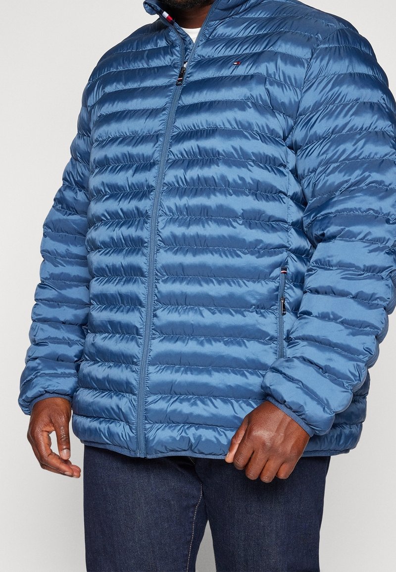 Veste doudoune bleue avec texture matelassée, fermeture éclair intégrale, col montant et deux poches latérales. Tissu lisse et brillant avec un logo de marque discret sur la poitrine.