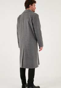 Uomo che indossa un lungo cappotto di lana grigio, pantaloni neri e scarpe nere, in piedi di spalle contro uno sfondo chiaro e uniforme.