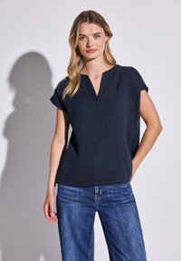 Marineblaue kurzärmlige Bluse mit V-Ausschnitt und strukturiertem Stoff. Getragen mit blauen Jeans, die eine lockere Passform und dezente Raffung aufweisen.