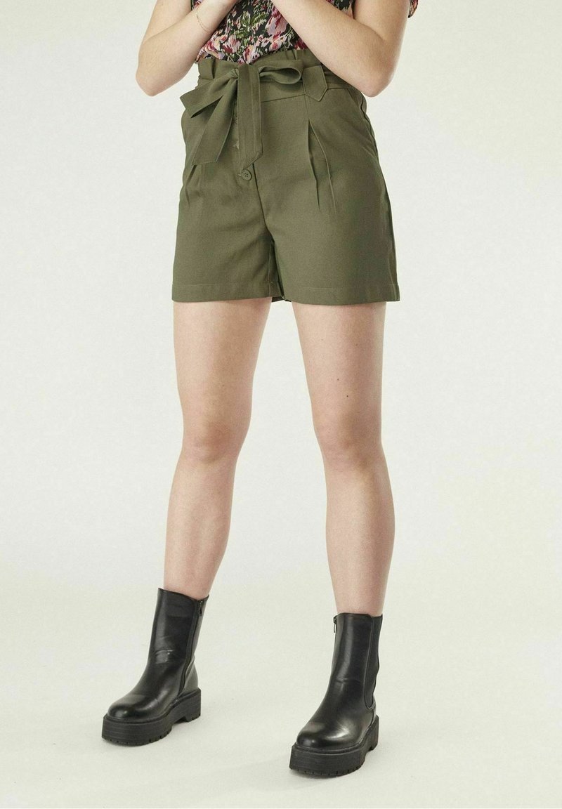 Personne portant un short taille haute vert olive avec une ceinture nouée et des bottines noires, debout devant un fond uni.
