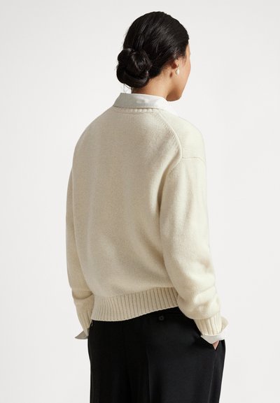 Kobieta z ciemnymi włosami związanymi w niski kok, ubrana w kremowy sweter na białej koszuli z kołnierzykiem oraz czarne spodnie, z rękami w kieszeniach, stoi tyłem.
