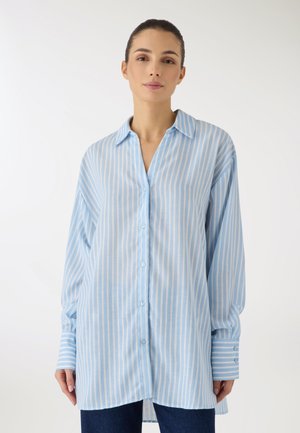 Donna che indossa una camicia a righe verticali lunghe azzurre e bianche, con bottoni e maniche lunghe, e pantaloni scuri, in piedi davanti a uno sfondo bianco.