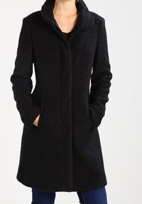 Donna che indossa un cappotto nero a trama, lungo fino al ginocchio, con un colletto alto, mani in tasca, abbinato a pantaloni blu scuro.
