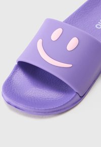 Lila csúszós szandál, texturált talppal és széles pánttal, rajta egy kiemelkedő rózsaszín mosolygós arc dizájnnal. Puha, gumiszerű anyagból készült.