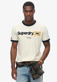 Camiseta de algodón de manga corta color crema con cuello y puños acanalados en navy, que presenta el logo "Superdry INC" en texto en negrita negro y naranja.