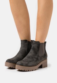 Czarne zamszowe botki za kostkę z grubymi brązowymi podeszwami i słupkowymi obcasami noszone na nagich nogach na tle białym.