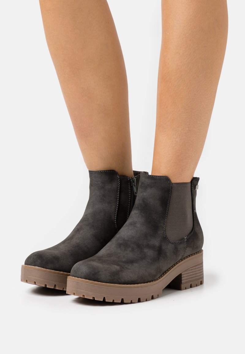 Czarne zamszowe botki za kostkę z grubymi brązowymi podeszwami i słupkowymi obcasami noszone na nagich nogach na tle białym.