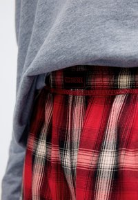 Röda och svarta rutig boxershorts med en svart "DIESEL"-etikett, delvis täckt av en grå, texturerad sweatshirt. Fokus på tyg- och tryckdetaljer.