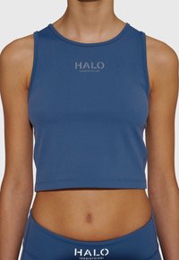 Kvinna som bär en blå ärmlös croptop och matchande leggings med "HALO"-logotyp centrerad på bröstet och midjebandet.