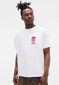 White cotton T-shirt featuring a red graphic print on the chest. Short sleeves and a round neckline. Soft texture with a relaxed fit.