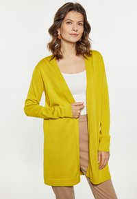Geel lange cardigan met een open voorkant, voorzien van lange mouwen en een zachte textuur. Gedragen over een witte tanktop en beige broek.
