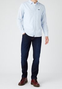 Camicia a righe azzurro chiaro con bottoni, jeans scuri e stivali marroni con lacci. La camicia ha una tasca sul petto e un colletto. Tessuto dalla texture liscia.