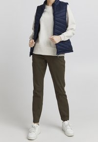 Tmavě modrá prošívaná vesta přes světle šedou mikinu, doplněná olivově zelenými kalhotami a bílými teniskami. Viditelné jsou zipy a elastické detaily.