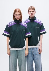 Πράσινο φούτερ με φερμουάρ, μοβ ώμοι με σχέδιο, λευκές λεπτομέρειες και λαστιχένιο τελείωμα. Φορεμένο με άνετο μπλε τζιν.