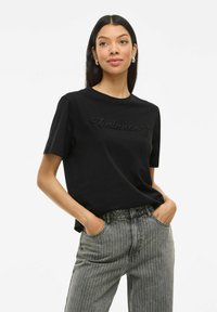 Svart bomullströja med "balance" broderat i matchande tråd, rund hals, parat med ljusgrå randiga jeans.