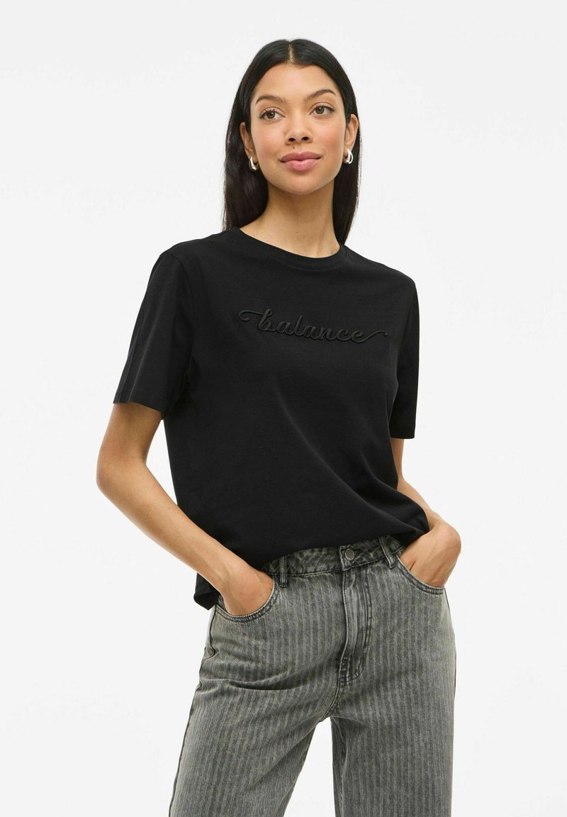 Svart bomullströja med "balance" broderat i matchande tråd, rund hals, parat med ljusgrå randiga jeans.