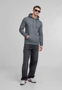 Felpa grigia con cappuccio e tasca frontale, abbinata a jeans scuri e sneakers bianche. Il modello indossa occhiali da sole scuri. Tessuto morbido.