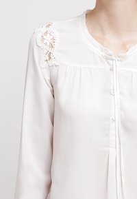 Blouse à manches longues, couleur crème pâle, avec des détails en dentelle sur les épaules, boutonnage à l'avant et plissé sur le devant. Tissu lisse avec une texture légère.