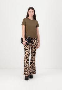 T-shirt olive green à manches courtes, associé à un pantalon beige à pattes évasées avec imprimé léopard et des chaussures à plateforme noires, tenant un sac à main noir avec une chaîne.