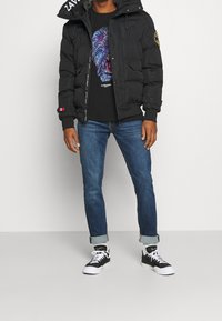 Person som bär en svart puffjacka, svart grafisk t-shirt, blå uppvikta jeans, vita strumpor och svart-vita sneakers stående på grått golv.