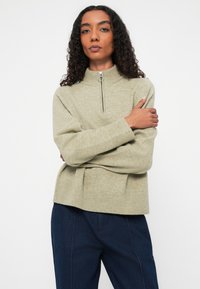 Suéter de punto verde claro con cuello alto y detalle de cremallera plateada. Presenta puños y dobladillo acanalados, acompañado de pantalones de mezclilla azul oscuro.